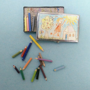 Een miniatuur kleurpotlodendoosje met een kindertekening op de deksel. Leuk detail: sommige kleurpotloodjes zijn aan het eind 'ingebeten'.
